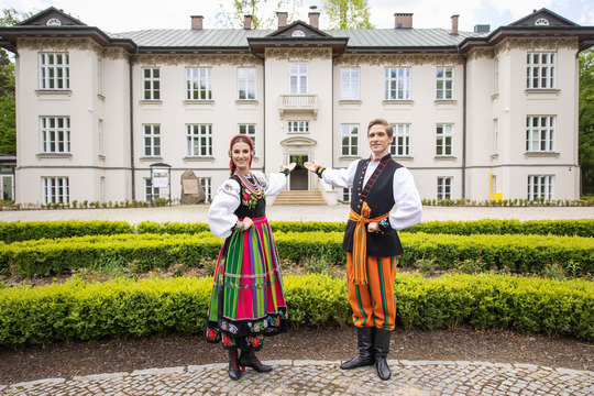 para taneczna z zespołu Mazowsze w kolorowych ludowych strojach pokazująca zabytkowy pałac, w którym mieści się Centrum Folkloru Polskiego „Karolin”